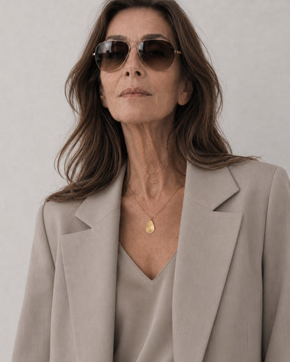 Time4Me gold chain necklace with a brushed teardrop pendant worn by a woman in a beige blazer and sunglasses.